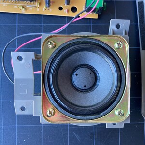 May include: A square-framed electronic speaker featuring a black circular paper cone with a central dust cap. The speaker is mounted on a gray plastic bracket with the label L1, secured by four gold-toned metal screws at each corner of the frame.