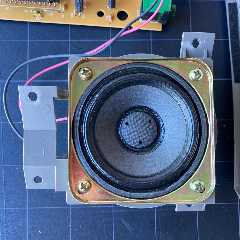 May include: A square-framed electronic speaker featuring a black circular paper cone with a central dust cap. The speaker is mounted on a gray plastic bracket with the label L1, secured by four gold-toned metal screws at each corner of the frame.
