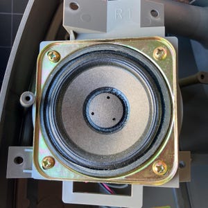 May include: A circular speaker driver mounted within a square, metallic, gold-toned frame featuring four corner screws. The speaker cone is dark gray with a black surround and a central dust cap marked with three small, evenly spaced holes. The text R1 is visible.