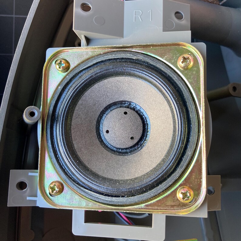 May include: A circular speaker driver mounted within a square, metallic, gold-toned frame featuring four corner screws. The speaker cone is dark gray with a black surround and a central dust cap marked with three small, evenly spaced holes. The text R1 is visible.