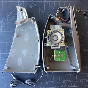 May include: A disassembled gray plastic speaker housing showing the internal components, including a circular speaker driver, a small green circuit board, wiring, and a black audio cable with a jack, alongside several small metal screws.