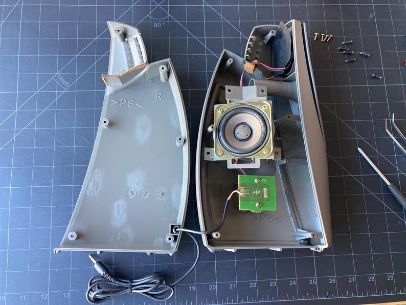 May include: A disassembled gray plastic speaker housing showing the internal components, including a circular speaker driver, a small green circuit board, wiring, and a black audio cable with a jack, alongside several small metal screws.
