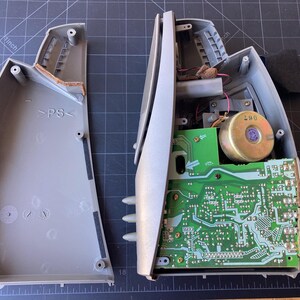 May include: A disassembled gray plastic speaker housing with its internal components exposed. The left side shows the empty interior shell with PS markings, while the right side displays a green circuit board, a circular speaker magnet, and wiring.