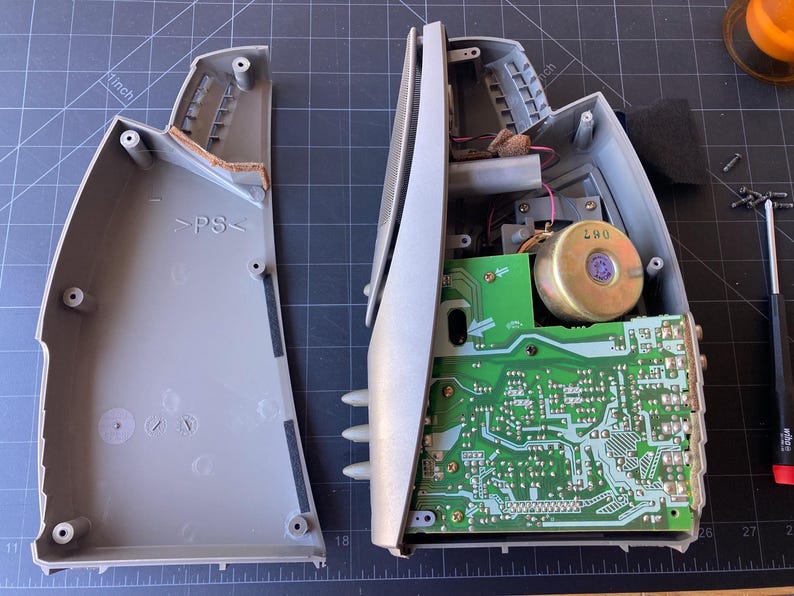 May include: A disassembled gray plastic speaker housing with its internal components exposed. The left side shows the empty interior shell with PS markings, while the right side displays a green circuit board, a circular speaker magnet, and wiring.