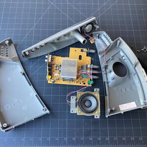 May include: A disassembled electronic device featuring a yellow circuit board, two gray plastic housing shells, a circular speaker, and several small metal screws. The internal components are exposed, showing wiring, a heat sink, and ports.