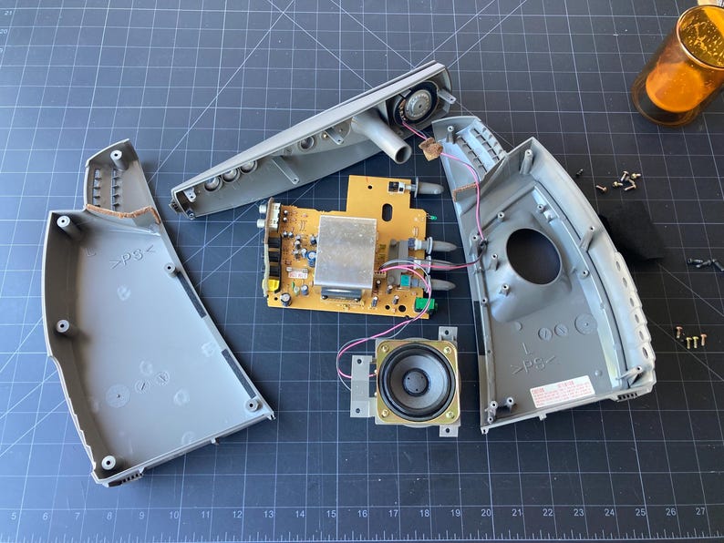 May include: A disassembled electronic device featuring a yellow circuit board, two gray plastic housing shells, a circular speaker, and several small metal screws. The internal components are exposed, showing wiring, a heat sink, and ports.