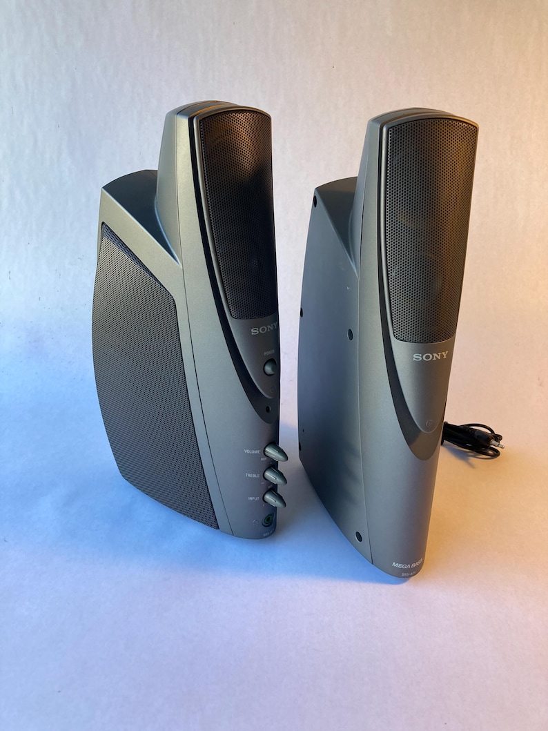 May include: A pair of dark gray Sony computer speakers with a vertical, curved design. The front features a black mesh grille, a power button, and three control knobs for volume, treble, and input. The base of the right speaker is labeled Mega Bass.