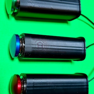 May include: Three cylindrical black devices, each featuring a metallic push-button cap on one end. The top cap is green, the middle cap is blue, and the bottom cap is red. Each device has a thin black wire extending from the opposite end.
