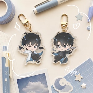 May include: Two acrylic keychains featuring chibi-style anime characters with black hair. One character wears a white coat and winks, while the other wears a dark coat and holds a sword. Both charms have gold-coloured lobster claw clasps.