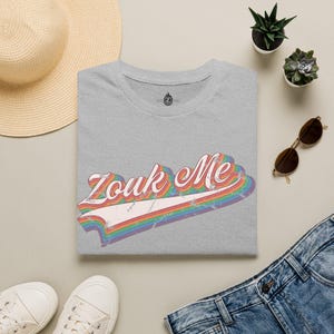 以下が含まれることがあります： ヘザーグレーのTシャツで、白くかすれたフォントで「Zouk Me」という文字が描かれ、その下にマルチカラーの虹色のストライプグラフィックが配置されています。リボンのような曲線的な形状と、使い古されたヴィンテージ風の質感が特徴のレトロなデザインです。