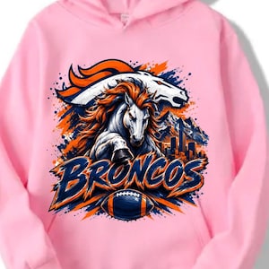 Puede incluir: Una sudadera con capucha de color rosa claro que presenta un estampado gráfico de un caballo blanco con crin naranja, una silueta de cabeza de caballo, un horizonte urbano y un balón de fútbol americano. El texto BRONCOS está en azul y naranja.