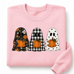 Könnte beinhalten: Ein hellrosa Sweatshirt mit Rundhalsausschnitt und einer Grafik von drei Geistern, die orangefarbene Kürbisse halten. Der erste Geist hat ein schwarzes Blumenmuster, der zweite ein schwarz-weißes Karomuster und der dritte ein Sternenmuster.