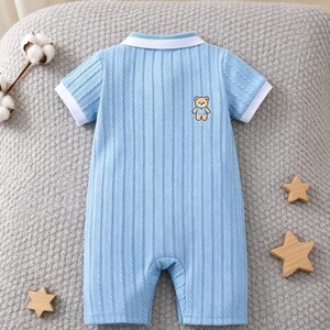 Op de afbeelding: Een lichtblauwe romper met korte mouwen, een verticale ribbelstructuur, een witte kraag en witte mouwboorden. Een kleine geborduurde bruine beer met een blauw shirt bevindt zich op het linkerbovenste gedeelte van de borst.
