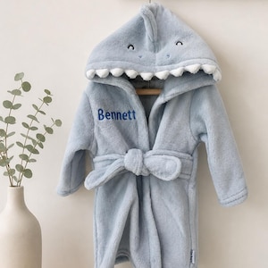May include: A light blue plush bathrobe featuring a hood designed like a shark with white teeth and embroidered eyes. The robe includes a matching tie belt and the name Bennett embroidered in dark blue thread on the upper left chest area.