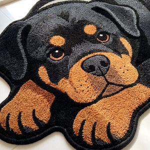 May include: A tufted rug shaped like a Rottweiler dog head and front paws. The rug features a black and tan color palette with a textured, plush surface. The dog has large brown eyes, a black nose, and tan markings above the eyes and on the muzzle.