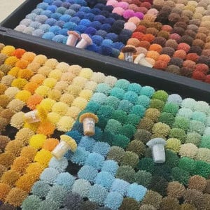 May include: A collection of small, round, fuzzy pom-poms arranged in a grid within a black tray. The pom-poms display a wide spectrum of colors, including shades of blue, green, yellow, orange, pink, and brown, with small numbered tags.