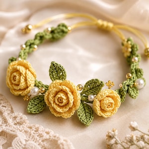 Op de afbeelding: Een gehaakte armband met drie gele rozen en groene bladeren, geaccentueerd met kleine witte parels en goudkleurige kralen. Het bloemmotief is gemaakt van garen, met een dun geel koord en een verstelbare schuifknoopsluiting voor een goede pasvorm.