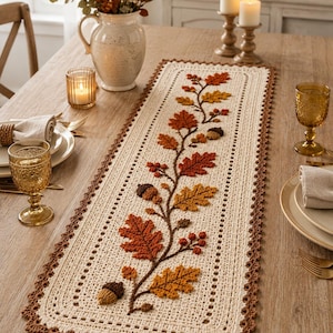 May include: A rectangular cream-colored crochet table runner featuring a central raised design of brown branches with textured orange, red, and yellow oak leaves and small brown acorns. The runner has a dark brown scalloped crochet border.