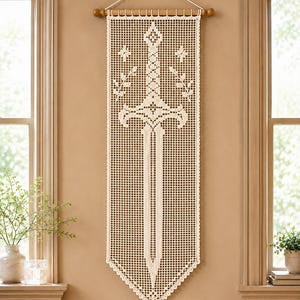 May include: A cream-coloured rectangular crochet wall hanging featuring a detailed sword design centred within a grid pattern. The piece is suspended from a wooden rod with a white cord and displays symmetrical leaf motifs on both sides of the sword.