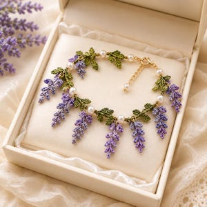 May include: A delicate bracelet featuring intricate crochet lavender sprigs in shades of purple and green, accented with small white pearls. The piece includes a gold-toned metal chain with a lobster clasp and an adjustable extender link section.