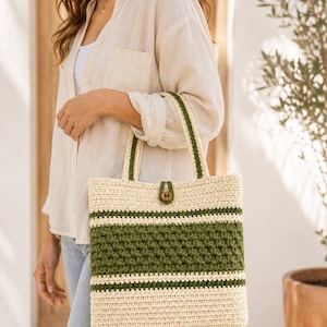 Puede incluir: Un bolso tote rectangular de ganchillo con rayas horizontales alternas en color crema y verde oliva. El bolso incluye dos asas de ganchillo a juego y una pequeña lengüeta de cierre central asegurada con un botón de madera marrón.