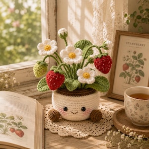 Peut inclure: Une plante fraise au crochet dans un pot blanc avec un visage souriant, des joues roses et des pieds marron. La plante présente des feuilles vertes, des fleurs blanches à cœur jaune, ainsi que des fraises rouges et vertes en laine.