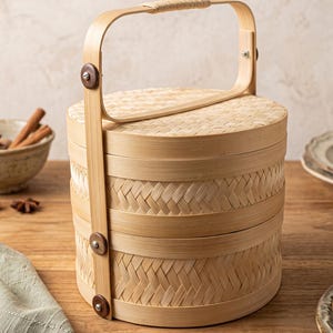 May include: A two-tiered round bamboo food container with a woven texture and a tall, arched handle. The structure is held together by vertical bamboo strips secured with dark circular fasteners, featuring a lid and two stackable compartments.