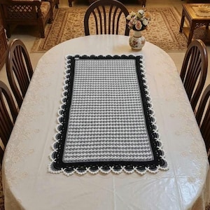 May include: A rectangular crochet table runner featuring a repeating geometric pattern in shades of gray and white. The runner is bordered by a thick black band and finished with a decorative white scalloped edge that frames the entire piece.