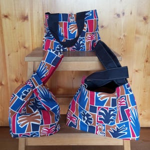 Op de afbeelding: Drie stoffen tassen met een geometrisch patroon in blauw, rood, bruin en wit. Eén tas heeft twee zwarte hengsels, terwijl de andere twee knooptassen zijn met één hengsel. Het patroon bevat gestileerde palmbomen en abstracte vormen.