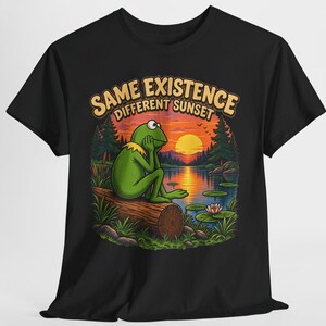 Könnte beinhalten: Ein schwarzes T-Shirt mit einer Grafik eines grünen Frosches, der bei Sonnenuntergang auf einem Baumstamm an einem See sitzt. Der Text über der Illustration lautet SAME EXISTENCE DIFFERENT SUNSET in heller Schrift. Das Motiv zeigt Bäume.