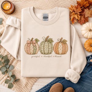 May include: A beige crewneck sweatshirt featuring a graphic of three pumpkins with varied patterns including floral, plaid, and solid textures. The text below the pumpkins reads grateful, thankful, blessed, separated by small heart icons.