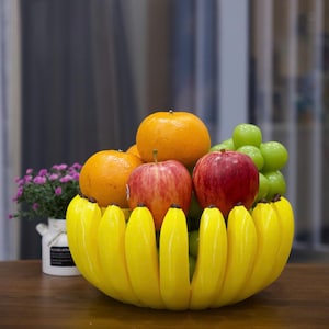 May include: A decorative fruit bowl shaped like a cluster of bright yellow bananas, filled with two red apples, two orange oranges, and a bunch of green grapes. The bowl is arranged with the banana peels forming the outer rim of the container.