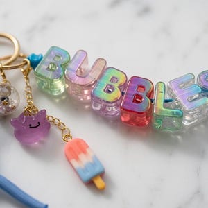 May include: A keychain featuring iridescent, multi-colored letter beads spelling BUBBLES, attached to a gold-toned metal ring. It includes a blue paracord loop, a small clear globe charm with tiny beads, a purple character charm, and a popsicle charm.