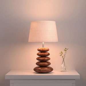 Puede incluir: Una l&aacute;mpara de mesa con una base formada por cinco formas ovaladas de madera marr&oacute;n apiladas y lisas. La l&aacute;mpara est&aacute; rematada con una pantalla de tela de color claro que emite un brillo suave y c&aacute;lido desde la fuente de luz interna.