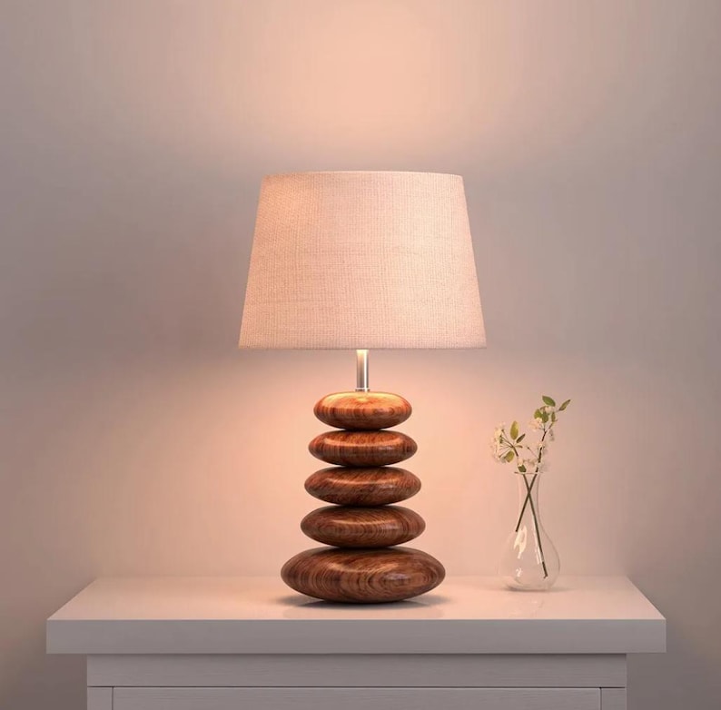 Puede incluir: Una l&aacute;mpara de mesa con una base formada por cinco formas ovaladas de madera marr&oacute;n apiladas y lisas. La l&aacute;mpara est&aacute; rematada con una pantalla de tela de color claro que emite un brillo suave y c&aacute;lido desde la fuente de luz interna.
