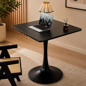 Puede incluir: Una mesa cuadrada negra con una base de pedestal única. Sobre la mesa hay un portátil plateado que muestra un paisaje urbano, una pequeña lámpara con pantalla beige plisada y un difusor de varillas de cristal con varillas negras.