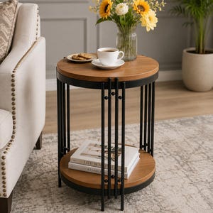 Puede incluir: Una mesa auxiliar redonda con dos estantes circulares de madera en acabado marrón cálido, sostenidos por una estructura de metal negro compuesta por varillas verticales. La mesa sostiene una taza de café blanca, un plato y un florero de cristal.