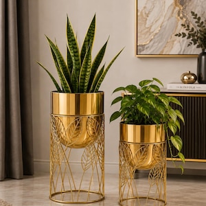 Op de afbeelding: Twee goudkleurige metalen plantenbakken met gladde cilindrische potten op gedetailleerde draadstandaards met een herhalend bladpatroon. De hogere bak bevat een sansevieria, de kortere bak bevat een hangende pothosplant.