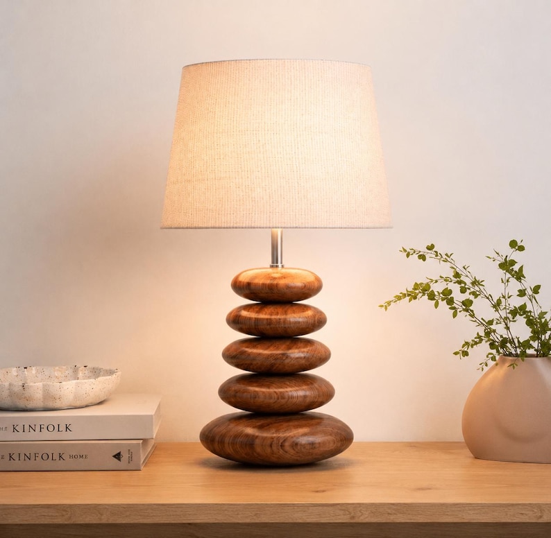 Puede incluir: Una l&aacute;mpara de mesa con una pantalla cil&iacute;ndrica de tela color hueso y una base formada por cinco piedras de madera pulida, lisas y apiladas, dispuestas en una columna vertical con una varilla met&aacute;lica que conecta la base con la luz.