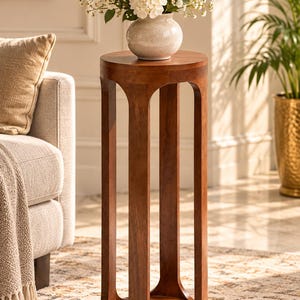 May include: A tall, cylindrical wooden plant stand featuring a smooth, medium-brown finish. The structure consists of a solid circular top and base connected by four vertical, arched supports that create an open, airy frame design.