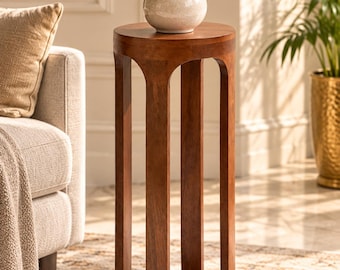 Puede incluir: Un soporte para plantas cilíndrico y alto de madera con un acabado liso en color marrón medio. La estructura consta de una parte superior circular sólida y una base conectadas por cuatro soportes verticales arqueados.