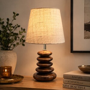 Puede incluir: Una l&aacute;mpara de mesa con una pantalla de tela beige texturizada y una base compuesta por cinco piedras de madera marr&oacute;n oscuro pulidas y apiladas. La l&aacute;mpara emite una luz c&aacute;lida que ilumina las formas de madera lisas y redondeadas.