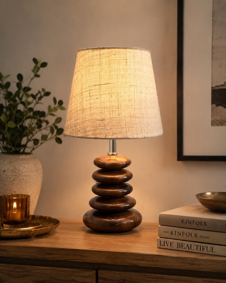 Puede incluir: Una l&aacute;mpara de mesa con una pantalla de tela beige texturizada y una base compuesta por cinco piedras de madera marr&oacute;n oscuro pulidas y apiladas. La l&aacute;mpara emite una luz c&aacute;lida que ilumina las formas de madera lisas y redondeadas.