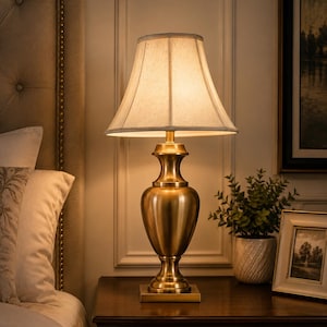May include: A table lamp with a metallic gold-toned urn-shaped base and a square pedestal. The lamp features a flared, off-white fabric lampshade that emits a warm, soft glow. The design includes a slender neck and a rounded body structure.