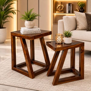 May include: Two dark brown wooden side tables with a geometric Z-shaped base design. The taller table holds a potted fern and two stacked books titled Kinfolk and Live Beautiful. The shorter table holds a gold-toned candle holder and a small succulent.