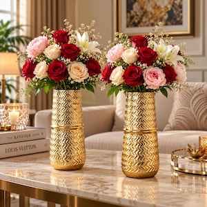 Può includere: Due vasi cilindrici dorati identici con un motivo a chevron testurizzato. Ogni vaso contiene una vivace composizione floreale con rose rosso scuro, peonie rosa tenue, rose color crema e gigli bianchi accentuati da piccoli rametti verdi.
