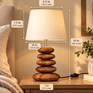 Puede incluir: Una l&aacute;mpara de mesa con pantalla de tela beige y base compuesta por cinco piedras apiladas con veteado de madera pulida. La l&aacute;mpara mide 48,3 cm de alto, con un di&aacute;metro superior de 16,5 cm, inferior de 25,4 cm y 17,8 cm de ancho de base.