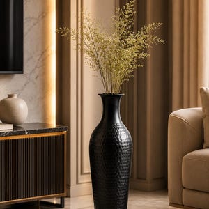May include: A tall, black floor vase with a textured, hammered surface and a narrow neck. The vase contains a cluster of delicate, light-colored dried grasses with thin, branching stems that extend upwards from the opening of the vessel.