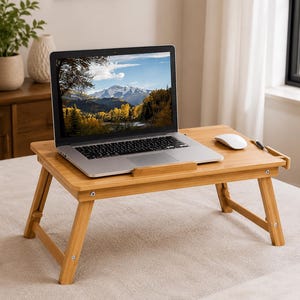 May include: A rectangular bamboo laptop desk with foldable legs, featuring a flat surface with a raised edge, a small side drawer, and a built-in stand for a laptop. A silver laptop with a mountain landscape screen display sits on the desk.