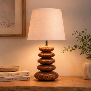 Puede incluir: Una l&aacute;mpara de mesa con una base compuesta por cinco piedras apiladas, pulidas y con textura de madera. La l&aacute;mpara tiene un cuello de metal plateado y una pantalla de tela c&oacute;nica de color blanco roto que emite una luz c&aacute;lida.